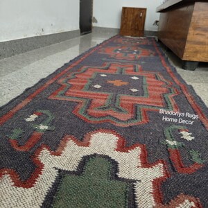 Puede incluir: Una alfombra larga y rectangular con un patr&oacute;n geom&eacute;trico en tonos azul marino, rojo, verde y blanco. La alfombra presenta un dise&ntilde;o central con formas repetidas y el texto "Bhadoriya Rugs Home Decor".