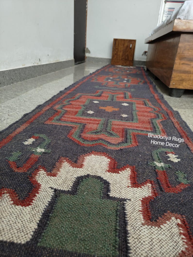 Puede incluir: Una alfombra larga y rectangular con un patr&oacute;n geom&eacute;trico en tonos azul marino, rojo, verde y blanco. La alfombra presenta un dise&ntilde;o central con formas repetidas y el texto "Bhadoriya Rugs Home Decor".