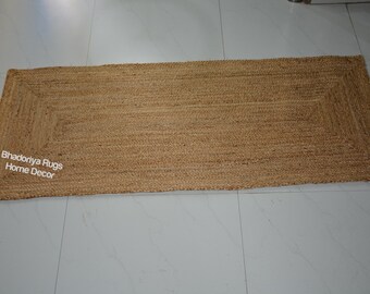 May include: A rectangular jute rug with a woven design. The rug is a natural tan color and has a rectangular border. The text "Bhadoriya Rugs Home Decor" is visible in the lower left corner. The rug is placed on a white tile floor.