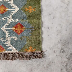 May include: A section of a woven rug with a geometric pattern. The rug features a blue-gray background with white, orange, and red diamond shapes. The border is green, and the rug has a fringe of brown tassels. The rug is on a gray surface.