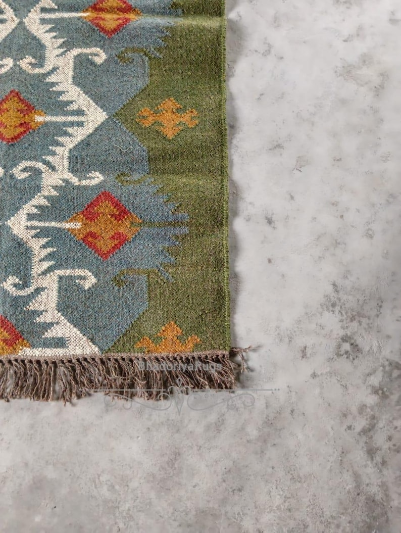 May include: A section of a woven rug with a geometric pattern. The rug features a blue-gray background with white, orange, and red diamond shapes. The border is green, and the rug has a fringe of brown tassels. The rug is on a gray surface.