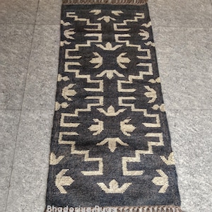 May include: A rectangular, dark gray rug with a geometric pattern in beige. The rug has a fringe on both ends and the text "Bhadoriya Rugs Home Decor" is visible at the bottom. The rug is placed on a gray tiled floor.
