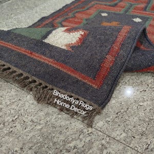 Puede incluir: Una alfombra rectangular enrollada con un patr&oacute;n geom&eacute;trico en tonos azul marino, rojo y verde. La alfombra tiene un borde con flecos y el texto "Bhadoriya Rugs Home Decor" es visible. La alfombra est&aacute; colocada sobre una superficie gris moteada.