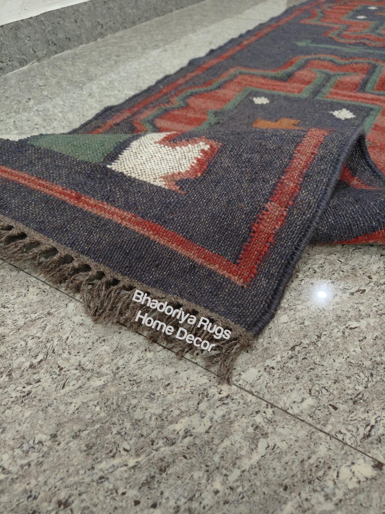Puede incluir: Una alfombra rectangular enrollada con un patr&oacute;n geom&eacute;trico en tonos azul marino, rojo y verde. La alfombra tiene un borde con flecos y el texto "Bhadoriya Rugs Home Decor" es visible. La alfombra est&aacute; colocada sobre una superficie gris moteada.