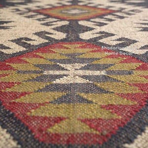 May include: A woven rug featuring a repeating geometric pattern in shades of cream, charcoal gray, olive green, and deep red. The textile displays a textured, coarse weave with visible fibers and the text Bhadoriya Home Decor printed on the surface.