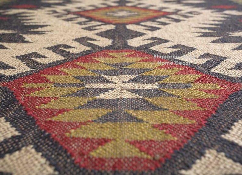 May include: A woven rug featuring a repeating geometric pattern in shades of cream, charcoal gray, olive green, and deep red. The textile displays a textured, coarse weave with visible fibers and the text Bhadoriya Home Decor printed on the surface.