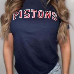 May include: Navy blue t-shirt with the word "PISTONS" in red, outlined in white. The shirt is a crew neck style and appears to be made of a soft material. The text is in a collegiate style font.