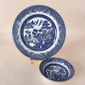 Op de afbeelding: Een blauw-wit Willow-patroon bord en kom set. Het bord heeft een gedetailleerde rand en een centrale scène van een pagode, brug en vogels. De kom weerspiegelt het ontwerp van het bord, waardoor een eetservies ontstaat.