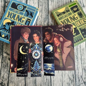 以下が含まれることがあります： イラストと3つのブックマークが付いた開いた本。本の表紙は青色で、「All The Young Dudes」というテキストが入っています。ブックマークには、天体と風景のデザインがあります。本の内部には人々のイラストが描かれています。