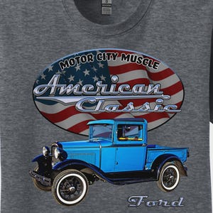 May include: Gray t-shirt featuring a blue vintage Ford pickup truck graphic. The design includes the text "Motor City Muscle" and "American Classic" over an American flag. The word "Ford" is also displayed.