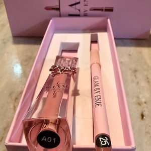 Puede incluir: Un set de maquillaje rosa en una caja rosa. El set incluye un brillo labial y un delineador de labios. El brillo labial está en un frasco transparente y angular con una tapa dorada. El delineador de labios es de color rosa claro con el número 25. La caja dice "IAM by Enie".