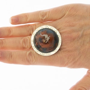 Op de afbeelding: Een zilveren ring met een ronde, bruine en zwarte harsinleg. De hars heeft een textuur en een organische uitstraling.