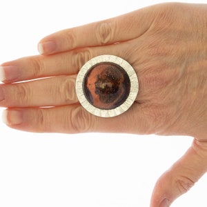 Op de afbeelding: Een zilveren ring met een grote, ronde, bruine hars steen gezet in een getextureerde zilveren bezel.