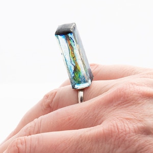 Op de afbeelding: Een zilveren ring met een rechthoekige blauwe, groene en zwarte hars steen.