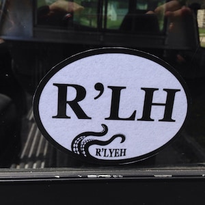 Peut inclure: Un autocollant ovale blanc avec du texte noir qui dit "R'LH" et un motif de tentacule noir avec le texte "R'LYEH" en dessous. L'autocollant est sur la vitre arrière d'un camion noir.