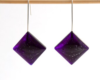 May include: A pair of dangle earrings with a square-shaped purple glass pendant. The glass has a sparkly, iridescent finish.