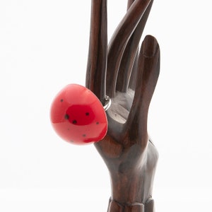Op de afbeelding: Een rode ring met zwarte stippen. De ring is gemaakt van keramiek en heeft de vorm van een koepel. De ring is op een houten hand.