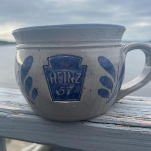 May include: A beige stoneware mug featuring a blue embossed Heinz 57 logo on the front. The mug has a rounded body, a wide rim, and a side handle. Blue leaf-like patterns are painted on both sides of the central logo, and the rim is accented with a blue band.