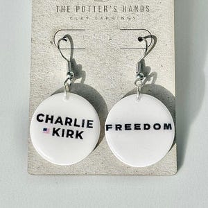 May include: White round earrings with the text "CHARLIE KIRK" and "FREEDOM" in black. The earrings are on a light gray card with the text "THE POTTER'S HANDS CLAY EARRINGS".