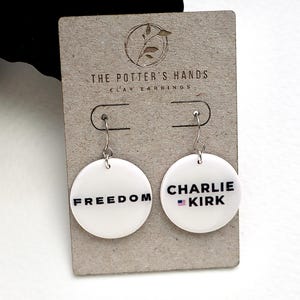 May include: White clay earrings with the words "FREEDOM" and "CHARLIE - KIRK" printed in black. The "CHARLIE - KIRK" earring has a small American flag graphic. The earrings are displayed on a gray card with the text "THE POTTER'S HANDS CLAY EARRINGS".