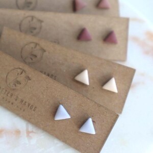 May include: Four pairs of triangle stud earrings in various colors: mauve, cream, and lavender. Each pair is displayed on a brown card with a logo. The earrings are small and geometric, perfect for everyday wear.
