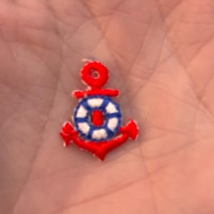 May include: A small, embroidered anchor patch in red, white, and blue. The anchor has a red body and a white and blue life preserver. The patch is likely for nautical-themed crafts or clothing.
