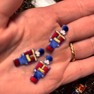 May include: Three small, embroidered patches in the shape of toy soldiers. The patches feature a blue uniform with red accents and a black hat. The patches are held in a hand.