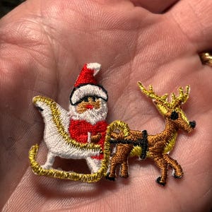 May include: Embroidered Christmas patch featuring Santa Claus in a sleigh pulled by a reindeer. Santa wears a red suit and hat, with a white beard. The sleigh is white with gold trim, and the reindeer is brown with yellow antlers.