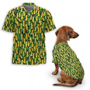 Op de afbeelding: Een button-down shirt met korte mouwen en een honden shirt, beide met een herhalend patroon van gele maïs op een zwarte achtergrond met groene stelen. Het honden shirt wordt gedragen door een bruine teckel. Het shirt heeft een klassieke kraag en knopen.
