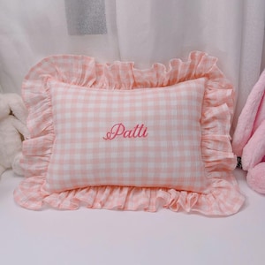 May include: A pink and white gingham pillow with a ruffled edge. The pillow has the name "Patti" embroidered in a cursive font. The pillow is rectangular and appears to be made of soft fabric.