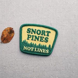 Könnte beinhalten: Gestickter Aufnäher mit dem Text "SNORT PINES NOT LINES" in Gelb und Grün. Der Aufnäher hat unten ein Walddesign und einen grünen Rand. Neben dem Aufnäher liegt ein getrocknetes Blatt.