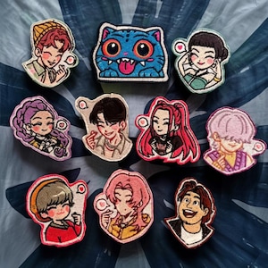 Peut inclure: Une collection d'écussons brodés avec des illustrations de style cartoon de personnes et d'un chat bleu. Les écussons sont de différentes couleurs, notamment le rouge, le rose et le violet. Chaque écusson a un design unique et est bordé d'un liseré blanc.
