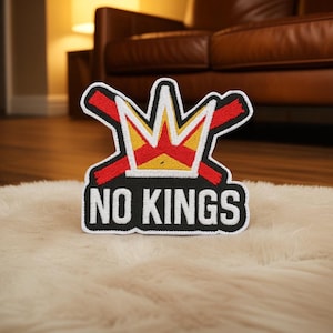 Op de afbeelding: Geborduurde patch met een kroonontwerp in rood, geel en wit, met de tekst "NO KINGS" in witte blokletters op een zwarte achtergrond. De patch heeft een witte rand en is bedoeld voor kleding of accessoires.