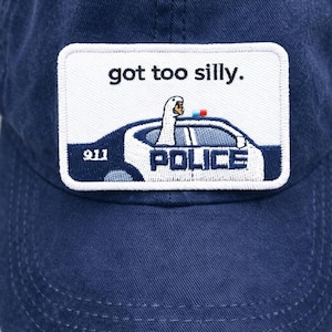 以下が含まれることがあります： 「got too silly」と書かれたパッチが付いた、濃いネイビーブルーの野球帽。パッチには、POLICEの文字と911の数字が入ったパトカーに乗った漫画のガチョウが描かれています。