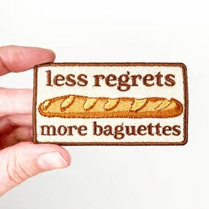 May include: Embroidered patch with a rectangular shape and a brown border. The patch features the text "less regrets more baguettes" in brown letters, with a detailed illustration of a baguette in the centre.