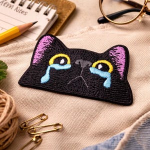 May include: Black embroidered cat patch with a crying face. The cat's eyes are yellow with blue tears, and the ears are purple. The patch is on a beige fabric surface.