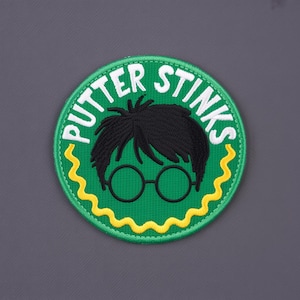 Könnte beinhalten: Ein runder, grüner Aufnäher mit dem Text "PUTTER STINKS" in Weiß. Der Aufnäher zeigt eine Cartoon-Illustration eines Charakters mit schwarzen Haaren, runden Gläsern und einem gelben Zickzack-Rand.
