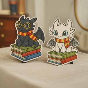 Może przedstawiać: Dwie haftowane naszywki z smokami. Jeden jest czarny z okularami i szalikiem, drugi biały. Oba smoki siedzą na stosach książek z czerwonymi, zielonymi i niebieskimi okładkami. Naszywki mają czarne obramowania.