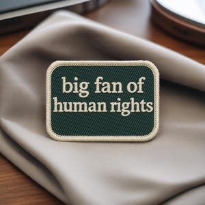 Könnte beinhalten: Ein rechteckiger Aufnäher mit abgerundeten Ecken, der den Text "big fan of human rights" in weißer Schrift auf dunkelgrünem Grund zeigt. Der Aufnäher hat einen cremefarbenen Rand und liegt auf einem neutralfarbenen Stoff.