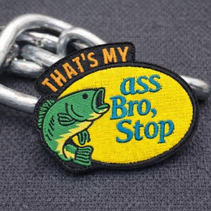 Pode incluir: Patch bordado com fundo oval amarelo e ilustração de peixe verde. O patch tem o texto "THAT'S MY ass Bro, Stop" em laranja e azul. O patch é colocado sobre uma superfície cinza texturizada com uma corrente prateada.