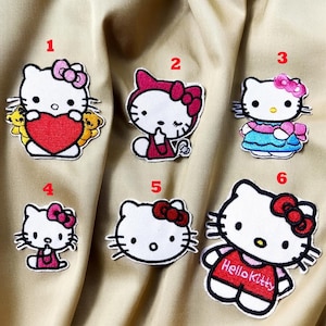 Peut inclure: Collection de six écussons Hello Kitty brodés. Chaque écusson représente le célèbre chat blanc avec un nœud rouge ou rose. Certains écussons incluent des cœurs, des tenues ou le texte "Hello Kitty". Les écussons sont sur un fond en tissu beige.