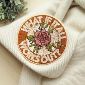 May include: Embroidered patch with the text "WHAT IF IT ALL WORKS OUT?" in a circular design. The patch features a central rose surrounded by white flowers and green leaves, set against a cream-colored background.