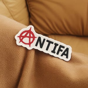 May include: A white patch featuring a red anarchist symbol and the black letters spelling "NTIFA". The patch is placed on a brown fabric, possibly part of a garment or accessory.
