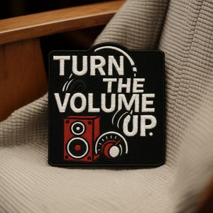 Può includere: Toppa quadrata nera con le parole "TURN THE VOLUME UP" in bianco. La toppa presenta un altoparlante rosso, cuffie e una manopola del volume. La toppa è su una superficie strutturata grigio chiaro.