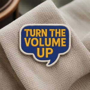 Può includere: Una toppa blu ricamata a forma di fumetto con le parole "TURN THE VOLUME UP" in giallo. La toppa ha un bordo bianco ed è posta su un tessuto di colore neutro.