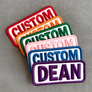 Peut inclure: Plusieurs écussons brodés de différentes couleurs : rouge, orange, vert, rose, bleu et violet. Chaque écusson présente un fond blanc avec le mot "CUSTOM" ou "DEAN" dans une couleur contrastante.