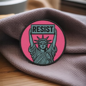 May include: A round patch featuring the Statue of Liberty holding a sign that reads "RESIST". The patch has a pink background with a black border. The Statue of Liberty is teal. The patch is on a purple fabric.
