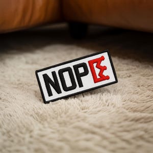 Könnte beinhalten: Ein rechteckiger Aufnäher mit dem Wort "NOPE" in schwarzen Großbuchstaben, wobei das "E" rot ist. Der Aufnäher hat einen weißen Hintergrund und einen schwarzen Rand. Der Aufnäher liegt auf einer flauschigen, hellfarbigen Oberfläche.