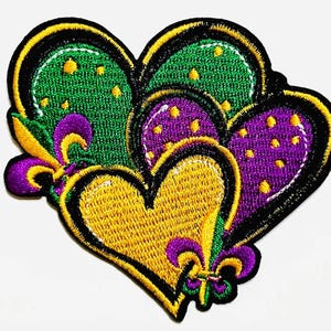 以下が含まれることがあります： 緑、紫、金色の3つの重なり合うハートが特徴の刺繍パッチ。パッチは黒い縁取りで、フルール・ド・リスのアクセントが付いています。ハートは小さな黄色の点で飾られています。