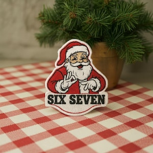 May include: Embroidered patch featuring Santa Claus in a red suit and hat, with a white beard and the text "SIX SEVEN". The patch is on a red and white checkered surface, with a small potted plant in the background.